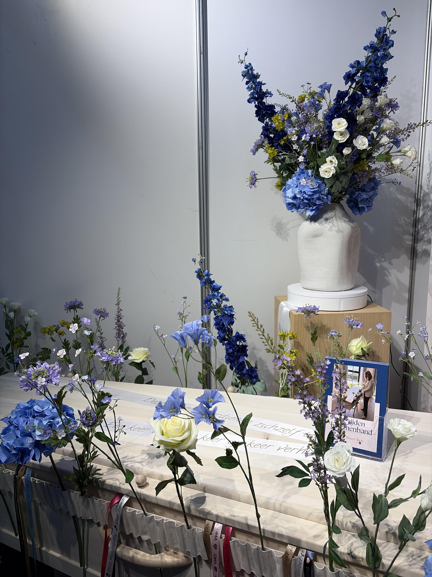 De beursstand van Flowers to Remember: kist met blauwe zijden bloemen en een witte vaas.